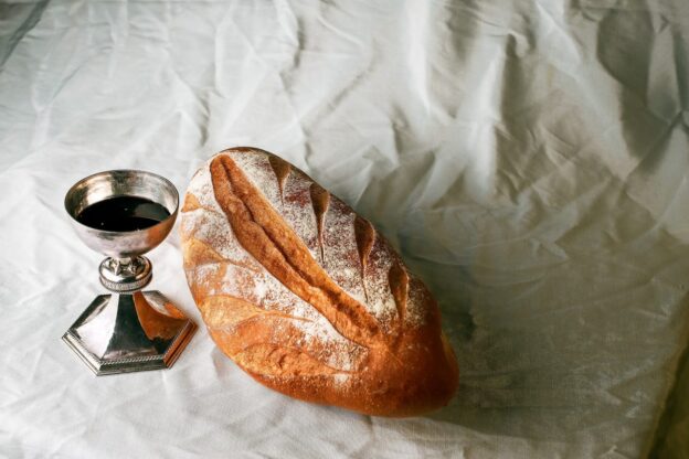 An image of holy communion featuring a silver cup of wine or juice and an artisanal loaf of bread dusted with flour and sitting on a white piece of cloth in subdued light and shadow.
