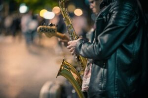 Musician playing saxophone on a city street at night with blurred lights in background.