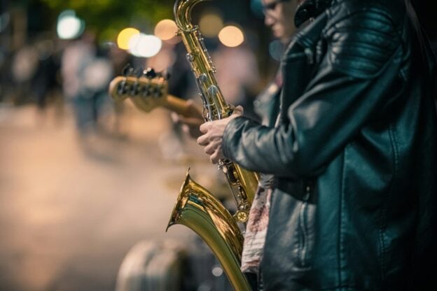 Musician playing saxophone on a city street at night with blurred lights in background.