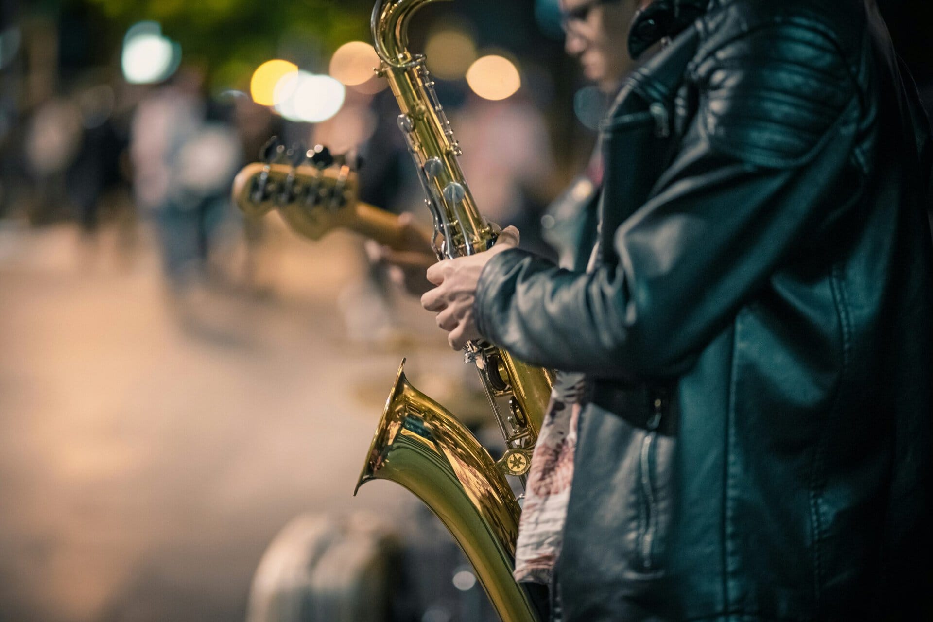 Musician playing saxophone on a city street at night with blurred lights in background.