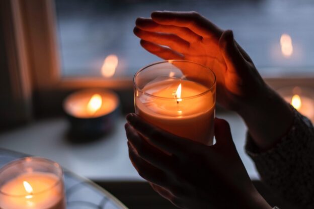 Hands cup around the warm glow of a candle in front of cool twilight shadows, flames of other candles flickering in the background
