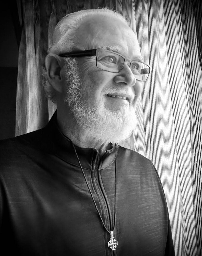 Elderly man with white hair and beard, wearing glasses and a cross necklace, smiling near curtains.