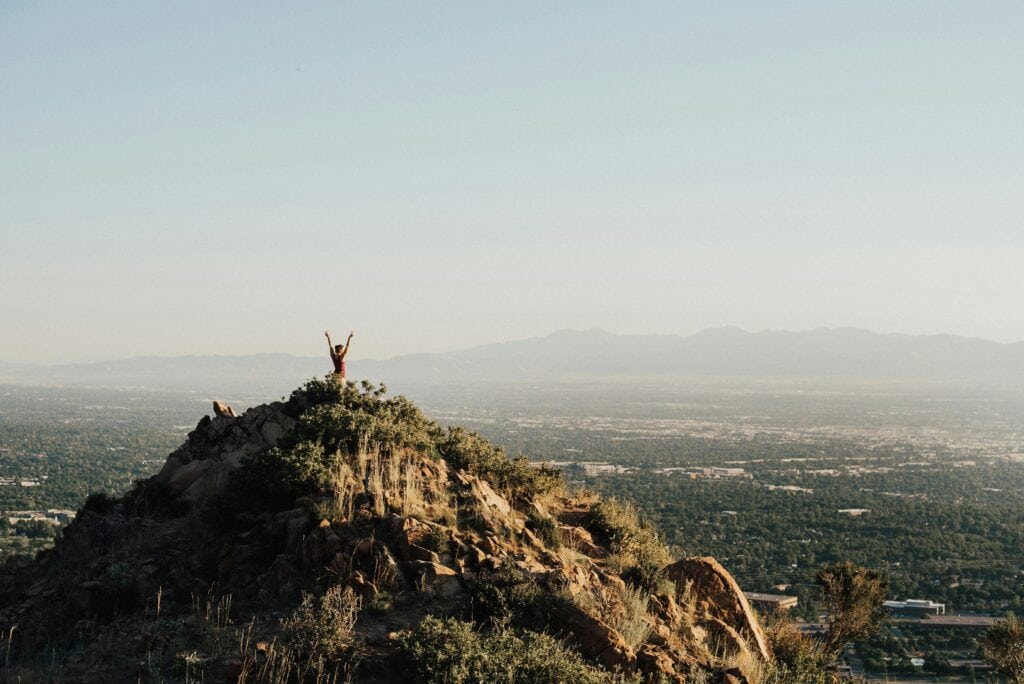 Person raising arms on mountain peak with city and mountains in background.