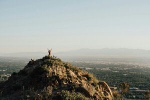 Person raising arms on mountain peak with city and mountains in background.