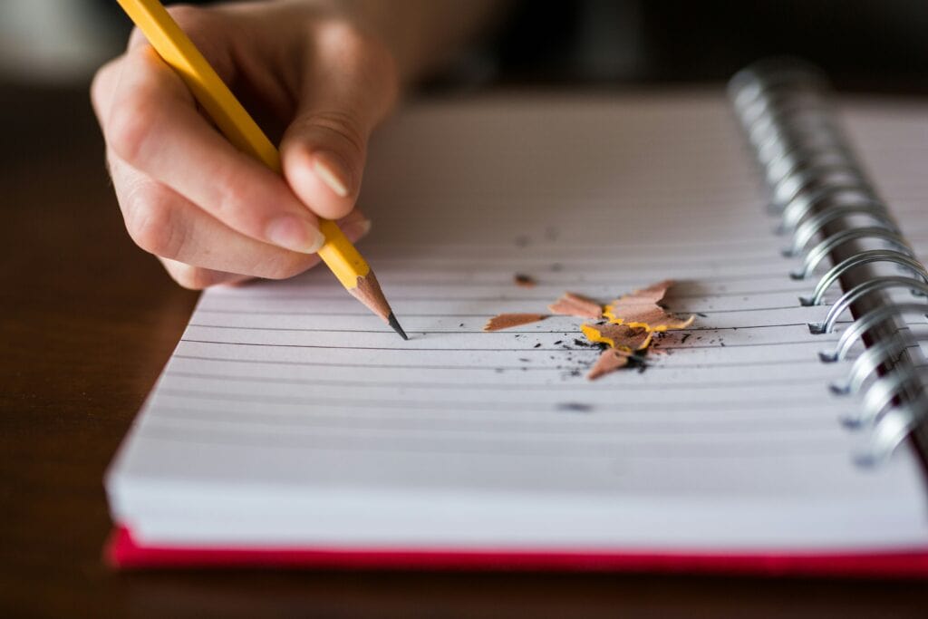 Person writing in a notebook with a pencil, with pencil shavings on paper.