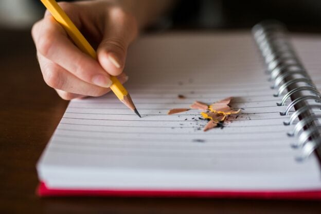 Person writing in a notebook with a pencil, with pencil shavings on paper.